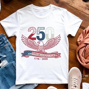 Op de afbeelding: Wit T-shirt met een afbeelding ter viering van de 250e verjaardag van de Amerikaanse Onafhankelijkheid. Het ontwerp toont een rood, wit en blauwe adelaar met uitgespreide vleugels, het nummer "250" en de tekst "American Independence 1776-2026".