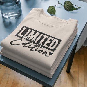 Könnte beinhalten: Ein Stapel gefalteter, cremefarbener T-Shirts. Die Shirts haben ein schwarzes rechteckiges Design mit der Aufschrift "LIMITED" über dem Wort "Edition" in Schreibschrift. Ein kleines schwarzes Herz befindet sich unter dem Wort "Edition". Die Shirts liegen auf einem hellblauen Tisch.
