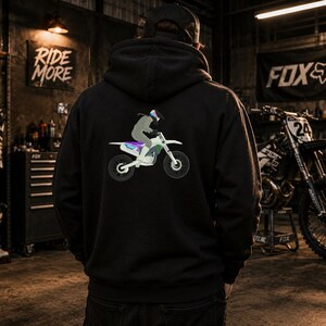 May include: Black hooded sweatshirt featuring a graphic of a person riding a dirt bike. The design includes a rider in gray and purple on a white and teal motorcycle. The background includes a motorcycle and the text "RIDE MORE".
