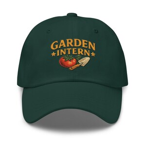 May include: A dark green baseball cap with the words "GARDEN INTERN" embroidered in gold, along with a tomato and trowel graphic. The cap has a curved brim and a button on top, suitable for gardening enthusiasts.