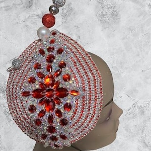 May include: A decorative headpiece with a teardrop shape, adorned with red and clear rhinestones. The headpiece features a central floral design with red gems and a beaded top with a pearl and red ball.