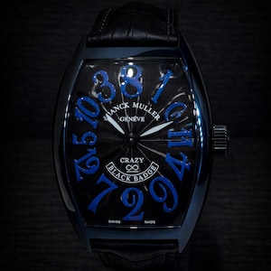 Könnte beinhalten: Schwarze Franck Muller Crazy Hours Uhr mit blauen Ziffern und Zeigern. Die Uhr hat ein schwarzes Lederarmband und ein schwarzes Zifferblatt mit den Worten "Franck Muller Geneve" und "Crazy Black Badge".