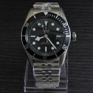 Puede incluir: Un reloj de pulsera de acero inoxidable con esfera y bisel negros. El reloj presenta una esfera negra con marcadores horarios luminosos, una ventana de fecha y la marca "TUDOR GENEVE". La pulsera es de acero inoxidable.