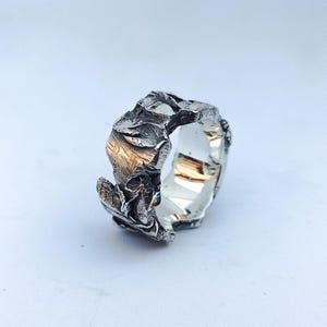 Puede incluir: Un anillo de plata con un diseño texturizado y robusto. El anillo tiene una forma irregular con una apariencia rugosa, similar a la piedra. El interior del anillo es liso y reflectante, contrastando con la superficie texturizada del exterior. El anillo está sobre un fondo blanco.