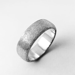 Puede incluir: Un anillo de tono plateado que presenta una superficie exterior texturizada y cepillada con un patrón lineal fino y una banda interior lisa y pulida. El metal tiene un acabado mate en la circunferencia exterior, contrastando con la superficie interior reflectante.