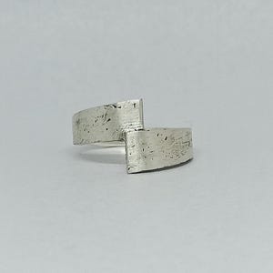 Puede incluir: Un anillo de plata con un diseño asimétrico único. El anillo presenta dos bandas curvas y texturizadas que se cruzan en ángulo recto. El metal tiene un acabado mate ligeramente rugoso, lo que le da una estética moderna y minimalista.