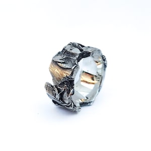 Puede incluir: Un anillo de banda ancha con un exterior de plata oxidada oscura que presenta una superficie rugosa, irregular y texturizada. El interior del anillo es liso y pulido, mostrando un acabado plateado brillante y reflectante con sutiles tonos dorados.