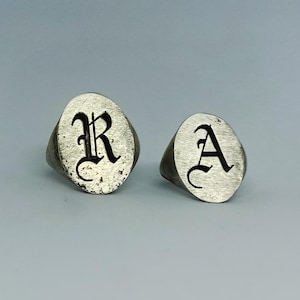 Puede incluir: Dos anillos de sello plateados con caras ovaladas. Un anillo presenta la letra "R" en fuente gótica negra, y el otro anillo muestra la letra "A" en un estilo similar. Los anillos tienen una apariencia texturizada y envejecida.