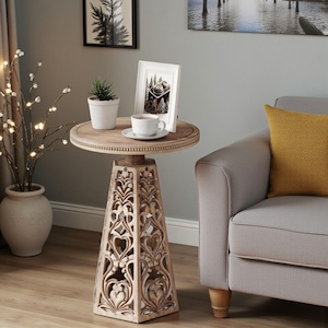May include: A round wooden side table with a light brown finish and a beaded edge on the tabletop. The base is a tall, tapered, four-sided structure featuring intricate, symmetrical carved cutouts with floral and heart-like patterns throughout.