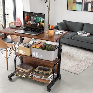 May include: A mobile rolling desk with a dark wood grain surface and black metal frame. The desk features a top tier with a laptop, a mug, and two storage bins labeled NO.2 and NO.3, and a lower shelf holding several stacked hardcover books.