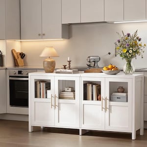 May include: A white kitchen sideboard with four glass-paneled doors and gold-toned handles. The top surface holds a beige table lamp, a stack of books, a candle holder, a bowl of yellow fruit, and a glass vase filled with colorful wildflowers.