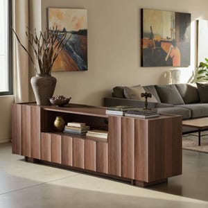 May include: A long, low-profile walnut wood media console featuring a fluted, vertical-slat design on the cabinet doors. The unit includes an open central shelving section containing stacked books and a small gold-toned decorative object.