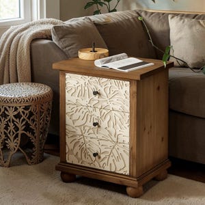 May include: A wooden side table with three drawers featuring a cream-colored leaf design. The table has a natural wood finish, black knobs, and rounded feet. A small wooden tray and an open magazine rest on top.