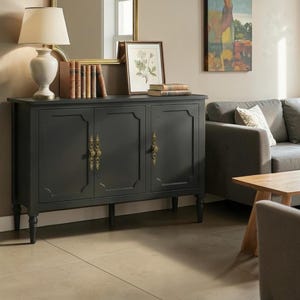 May include: A charcoal gray sideboard featuring three cabinet doors with ornate gold-toned metal handles. The piece stands on four turned legs and has a flat top surface holding a white table lamp, a row of vintage books, and a framed botanical print.
