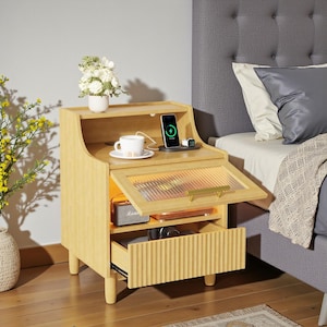 May include: A light wood bedside table with a drawer and a cabinet with a glass door. A phone is charging on top, next to a cup and a small white vase with flowers. A small speaker is inside the cabinet.