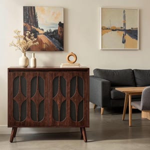 May include: A dark brown wooden cabinet featuring two doors with four vertical oval cutouts and dark mesh panels. The cabinet stands on four tapered legs and is topped with two cream-colored ceramic vases, a small circular wooden sculpture, and books.