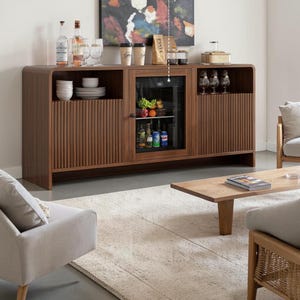 May include: A dark wood sideboard featuring a central glass-fronted beverage refrigerator, flanked by two cabinets with vertical fluted detailing. The unit includes open shelving on the left and right sides, providing storage for glassware, plates, and bottles.