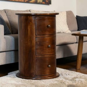 May include: A dark brown, cylindrical wooden nightstand with three drawers. Each drawer has a small, round, silver-colored knob. The nightstand sits on a round base and has a rounded top. The wood grain is visible.