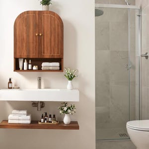 K&ouml;nnte beinhalten: Ein wandmontierter Badezimmerschrank aus Holz mit gew&ouml;lbtem Oberteil und zwei T&uuml;ren, verziert mit vertikalen Einlegearbeiten und zwei kleinen goldenen Kn&ouml;pfen. Darunter befinden sich ein wei&szlig;es rechteckiges Waschbecken, ein Holzregal und wei&szlig;e Handt&uuml;cher.