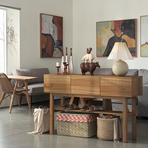 May include: A wooden console table with three drawers and a lower shelf, featuring a table lamp with a pleated shade, animal figurines, and decorative candlesticks. Below, a woven basket, a patterned storage box, and a fabric bag are placed.