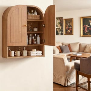 K&ouml;nnte beinhalten: Ein h&auml;ngender Holzschrank mit gew&ouml;lbtem Oberteil und zwei T&uuml;ren, von denen eine ge&ouml;ffnet ist. Das Innere verf&uuml;gt &uuml;ber drei Regale mit verschiedenen Flaschen, einem geflochtenen Korb und einem Duftdiffusor. Ein offenes unteres Regal enth&auml;lt zwei Becher, einen Seifenspender und gefaltete Handt&uuml;cher.