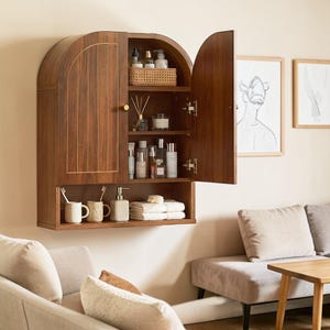 K&ouml;nnte beinhalten: Ein wandmontierter Schrank aus dunklem Holz mit gew&ouml;lbter Oberseite und zwei T&uuml;ren. Das Innere verf&uuml;gt &uuml;ber drei Regale mit verschiedenen Flaschen, einem geflochtenen Korb und einem Duftdiffusor. Unter den T&uuml;ren befindet sich ein offenes Regal mit zwei Bechern, einem Seifenspender und gefalteten wei&szlig;en Handt&uuml;chern.