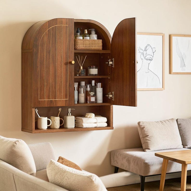 K&ouml;nnte beinhalten: Ein wandmontierter Schrank aus dunklem Holz mit gew&ouml;lbter Oberseite und zwei T&uuml;ren. Das Innere verf&uuml;gt &uuml;ber drei Regale mit verschiedenen Flaschen, einem geflochtenen Korb und einem Duftdiffusor. Unter den T&uuml;ren befindet sich ein offenes Regal mit zwei Bechern, einem Seifenspender und gefalteten wei&szlig;en Handt&uuml;chern.