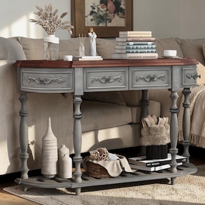 May include: A gray and brown console table with two drawers, ornate carvings, and turned legs. The table has a lower shelf and is decorated with vases, books, and a decorative basket. The table is placed in front of a beige sofa.