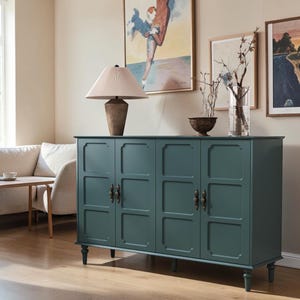 May include: A dark teal four-door sideboard cabinet featuring recessed rectangular panels on each door, antique brass-toned hardware, and turned legs. The top surface holds a table lamp with a pleated shade and a clear glass vase with branches.