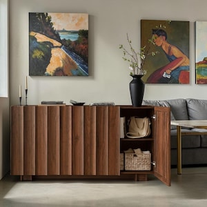 May include: A dark brown wooden sideboard featuring a vertical slatted front design. The unit has one open cabinet section revealing two interior shelves, one containing a beige handbag and the other a woven basket with light-colored items.