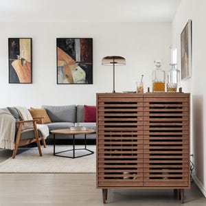 May include: A rectangular wooden cabinet with horizontal slatted doors, standing on four tapered legs. The cabinet features a grid-like interior structure for storage and is topped with a modern lamp and two glass decanters filled with amber liquid.