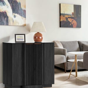 May include: A black wooden sideboard with a fluted center panel design and two side doors. A round, glossy, burnt-orange ceramic table lamp with a beige fabric shade sits on top, next to a small, rectangular, black-framed picture frame.