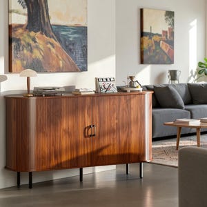 May include: A long, oval-shaped wooden sideboard featuring a vertical slatted texture, two central doors with black metal handles, and four slender black cylindrical legs. The top surface holds a small lamp, a record player, and a book display.