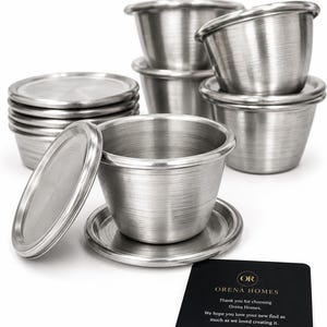 Pode incluir: Um conjunto de tigelas de servir em metal escovado com tampas e pires a condizer. As tigelas são empilhadas e dispostas, com uma tigela e tampa separadas. Um cartão preto com texto dourado diz "ORENA HOMES".