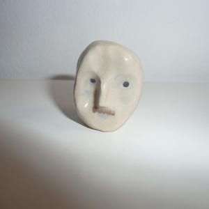 May include: A small, off-white ceramic sculpture shaped like a human face. The piece features a prominent nose, two dark circular eyes, and a faint brown mustache detail. The surface has a smooth, glossy finish with subtle speckling throughout.
