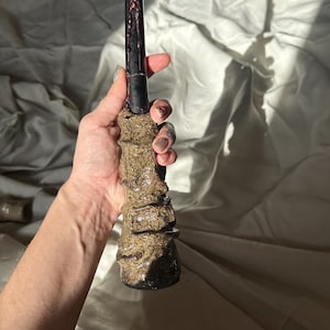 Peut inclure: Une bougie noire avec des coulures de cire rouge, tenue par une main. La bougie repose sur une base marron texturée. Un objet décoratif unique.
