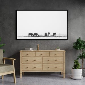 May include: Monochrome framed print depicting horses in a snowy field with a person. The artwork hangs above a light wood chest of drawers with seven drawers. A chair, plants, and decor are in the foreground.