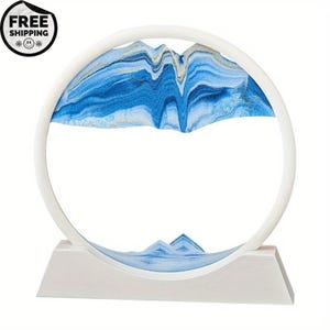 May include: A white, circular sand art piece with a rectangular base. The sand art features blue and white sand, creating a mountain-like landscape within the circle. The piece is designed to be rotated, allowing the sand to shift and create new patterns.