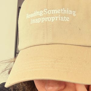 May include: Tan baseball cap with the embroidered text "Reading Something Inappropriate". The cap has a curved brim and a classic six-panel design. The hat is made of a soft, durable fabric.