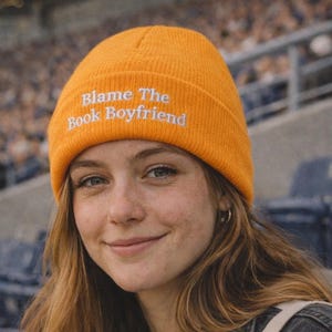 May include: An orange beanie with the text "Blame The Book Boyfriend" embroidered in white. The person is wearing a grey top and a black denim jacket. They are holding a coffee cup with a red straw.