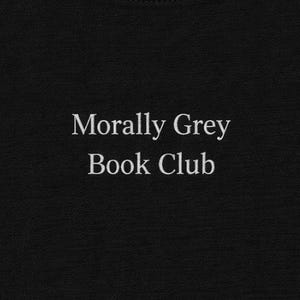 Peut inclure: T-shirt noir avec l'inscription blanche "Morally Grey Book Club". Le texte est centré. Le tissu sombre suggère un vêtement confortable et décontracté.