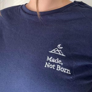 May include: A navy blue t-shirt featuring a white mountain and moon design. The words "Made, Not Born" are printed beneath the graphic. The top has a crew neck and short sleeves.