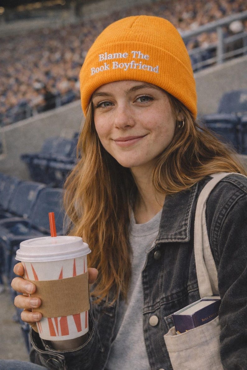 May include: An orange beanie with the text "Blame The Book Boyfriend" embroidered in white. The person is wearing a grey top and a black denim jacket. They are holding a coffee cup with a red straw.