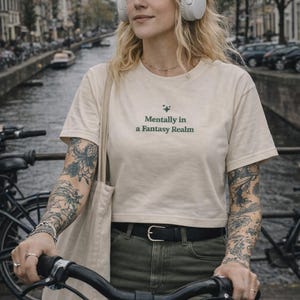 K&ouml;nnte beinhalten: Cremefarbenes Crop-Top mit dem Text "Mentally in a Fantasy Realm" in Gr&uuml;n. Die Person tr&auml;gt wei&szlig;e Kopfh&ouml;rer und f&auml;hrt Fahrrad. Die Person tr&auml;gt einen schwarzen G&uuml;rtel und olivgr&uuml;ne Hosen. Der Hintergrund zeigt einen Kanal und Geb&auml;ude.