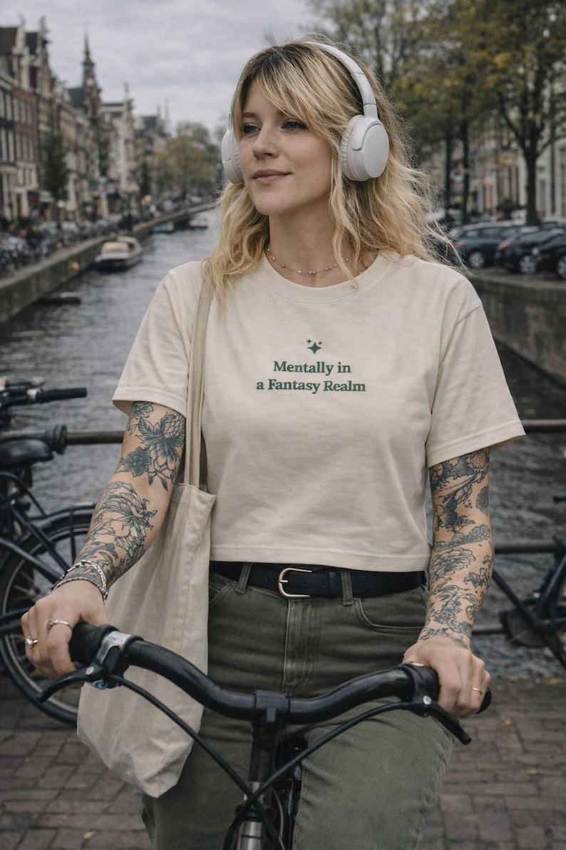 K&ouml;nnte beinhalten: Cremefarbenes Crop-Top mit dem Text "Mentally in a Fantasy Realm" in Gr&uuml;n. Die Person tr&auml;gt wei&szlig;e Kopfh&ouml;rer und f&auml;hrt Fahrrad. Die Person tr&auml;gt einen schwarzen G&uuml;rtel und olivgr&uuml;ne Hosen. Der Hintergrund zeigt einen Kanal und Geb&auml;ude.