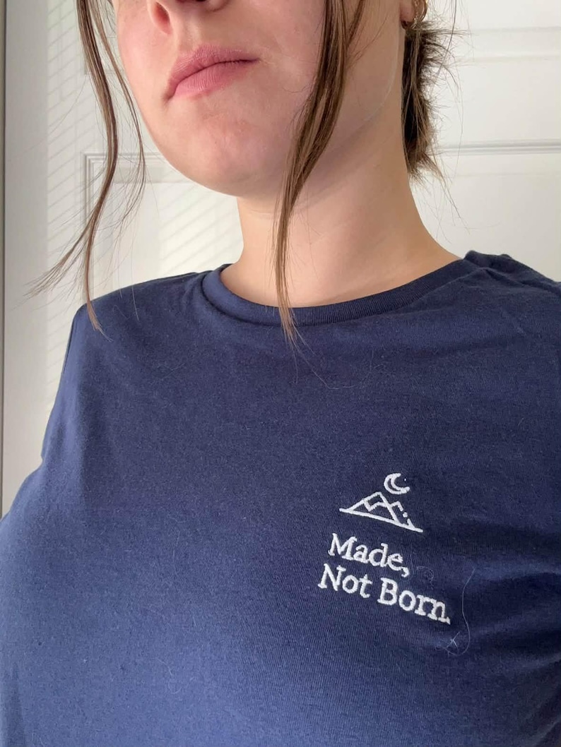 May include: A navy blue t-shirt featuring a white mountain and moon design. The words "Made, Not Born" are printed beneath the graphic. The top has a crew neck and short sleeves.
