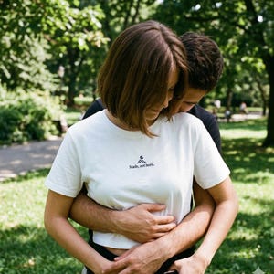 May include: A white t-shirt with the text "Made, not born." is worn by a person. The shirt has a small graphic above the text. The person is embraced by another person, with a green park background.