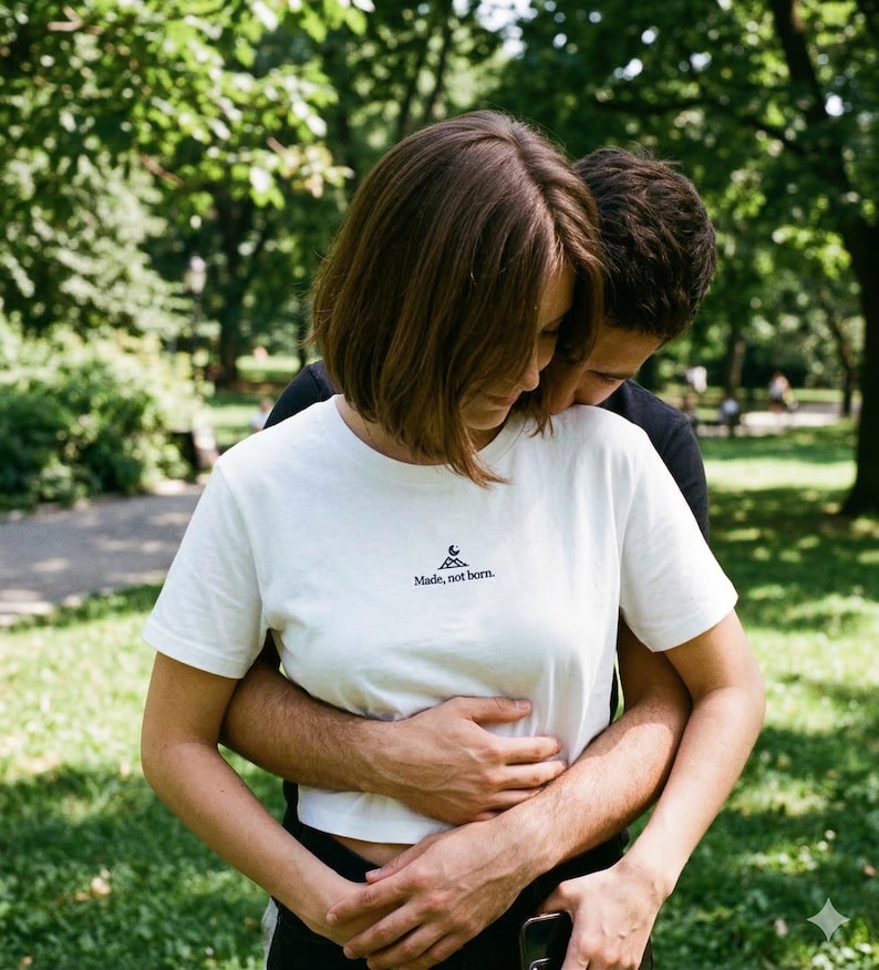May include: A white t-shirt with the text "Made, not born." is worn by a person. The shirt has a small graphic above the text. The person is embraced by another person, with a green park background.