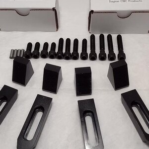 May include: A collection of black metal clamps, bolts, and wedges, likely for industrial use. The components are arranged on a white surface, with two white cardboard boxes in the background. The items appear new and unused.