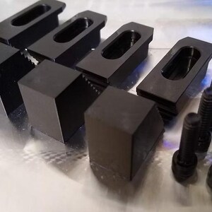 May include: A collection of black metal clamps and bolts. The clamps have a rectangular base and a slotted top. The bolts are cylindrical with a hex head. The items are arranged on a reflective surface.